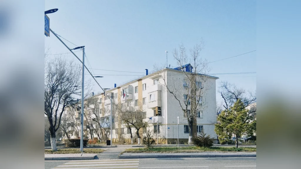 A residential street in Almaty with private houses and overhead power lines, with a local resident on the sidewalk. Documentary photograph in natural daylight, with no text.