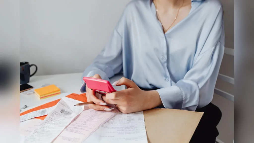 A woman plans her household budget with a laptop and notebook at a kitchen table in an Almaty apartment. An urban residential neighbourhood is visible through the window. Natural light, with no text o