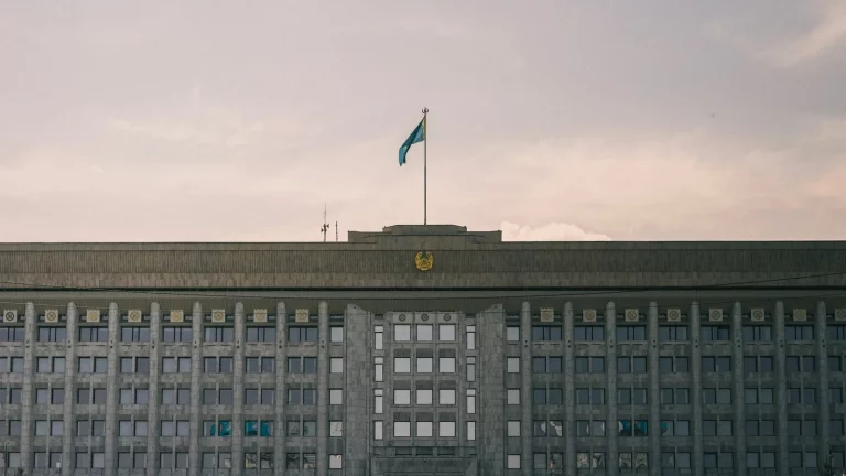 Қазақстандағы аумақтық әділет органы ғимаратының жанындағы деректі кадр: қолына құжат салынған папка ұстаған адам кіреберіске жақындап келеді. Мәтін мен оқуға болатын дербес деректер жоқ.