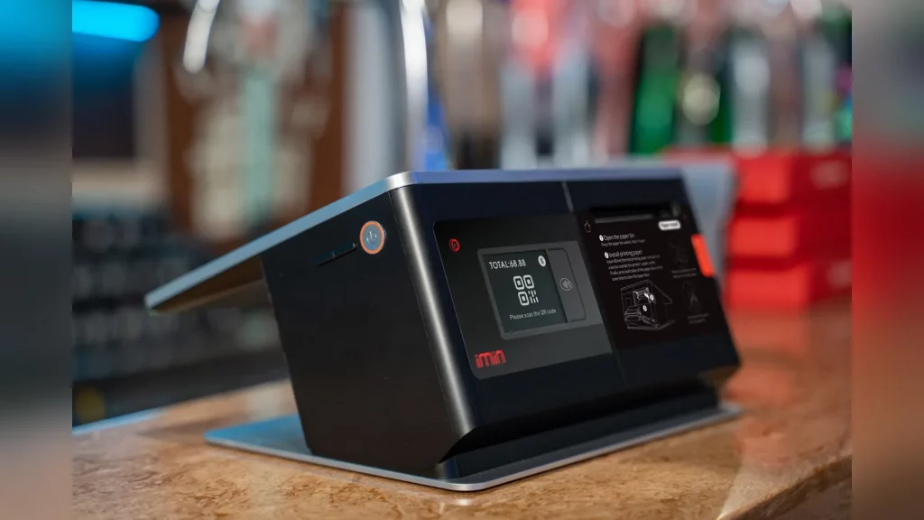 Владелец небольшого магазина в Казахстане принимает оплату по QR-коду. Покупатель держит смартфон у кассы, на фоне видны товары и рабочее место продавца. Документальная фотография, естественный свет,