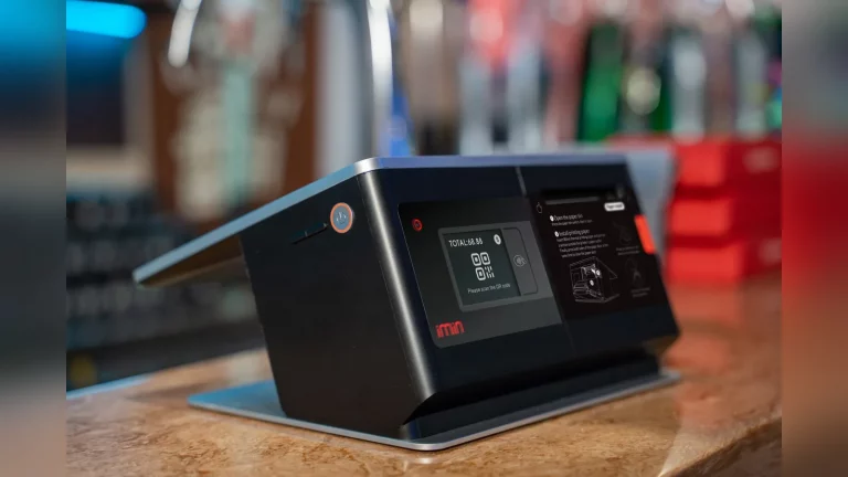 Қазақстандағы шағын дүкен иесі QR-код арқылы төлем қабылдап жатыр. Сатып алушы касса жанында смартфон ұстап тұр, артқы жақта тауарлар мен сатушының жұмыс орны көрінеді. Табиғи жарықта түсірілген дерек