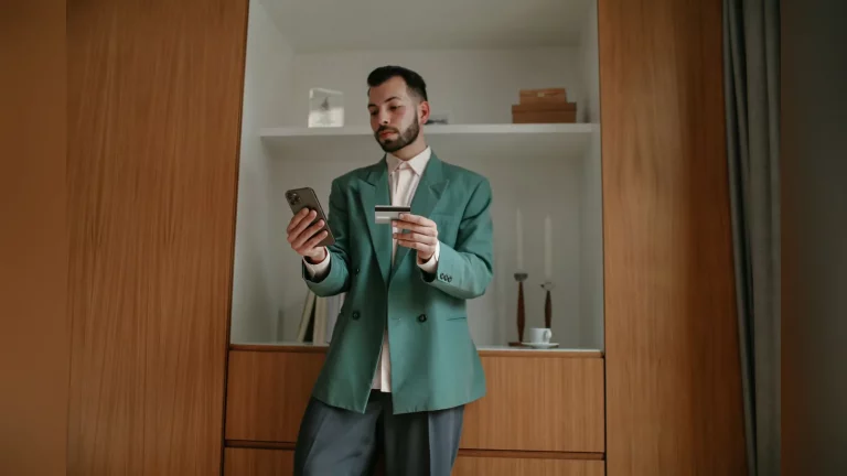 Күзде қала көшесіндегі Halyk бөлімшесінің жанында смартфон ұстап тұрған Алматы тұрғыны. Табиғи күнделікті көрініс, мәтін де, экранда оқылатын деректер де жоқ.
