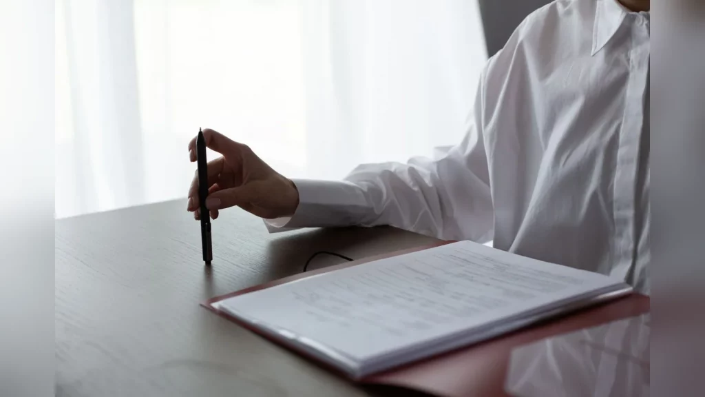 Candid photograph in a small business office in Kazakhstan: an employee compares work documents on a laptop, with a colleague holding a folder nearby. Natural light, with no text or readable data on t