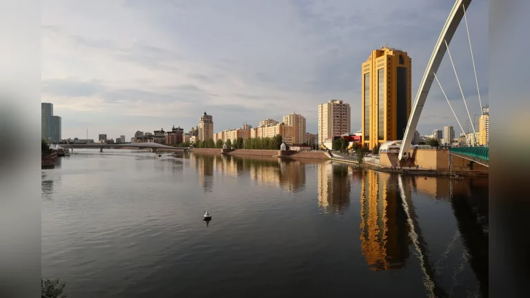 Documentary photograph of infrastructure construction in Kazakhstan, with a worker in the foreground and construction machinery and a site being upgraded in the background. No text or graphics.