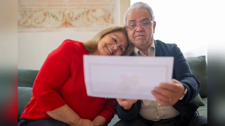 A man and woman aged around 55 sit at a table in a bright apartment in Kazakhstan, discussing documents and looking at a laptop. Natural daylight, a calm everyday setting, with no text or logos.